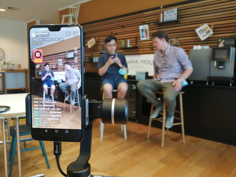 Blick auf den Live-Stream über ein Smartphone-Display. Im Hintergrund: Hanno Rademacher und sein Co-Moderator sitzen im Hotel Vienna House Easy.